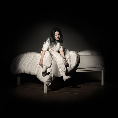 American singer-songwriter Billie Eilish sits on the edge of a messy white-sheeted bed in a dark, shadowy room. She wears an all-white t-shirt and pants, leaning forward with a wide, manic grin. Her eyes are entirely white and pupil-less, glowing under a single spotlight that creates a stark contrast against the deep black background.